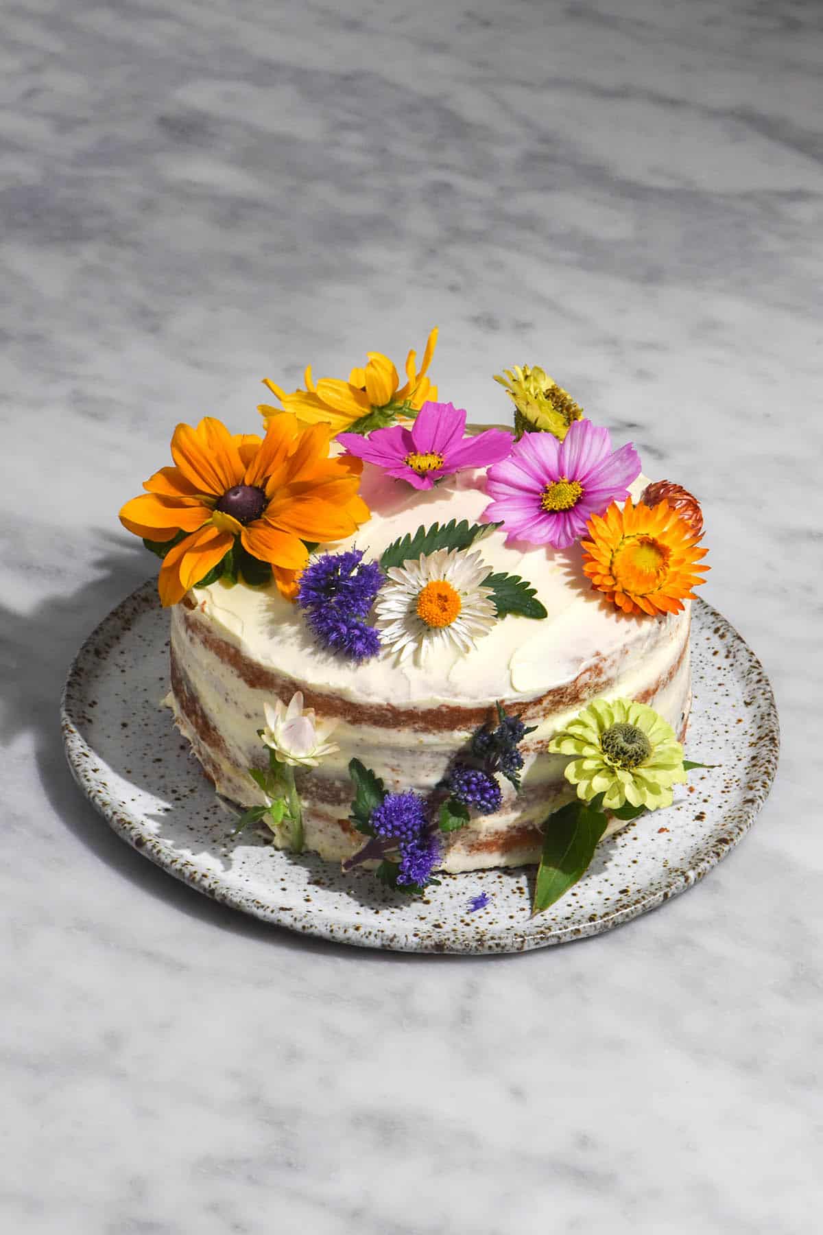 An aerial image of a gluten free cake with passionfruit curd filling and cream cheese icing. The cake is decorated with multi coloured flowers and sits on a white speckled ceramic plate atop a white marble table. 