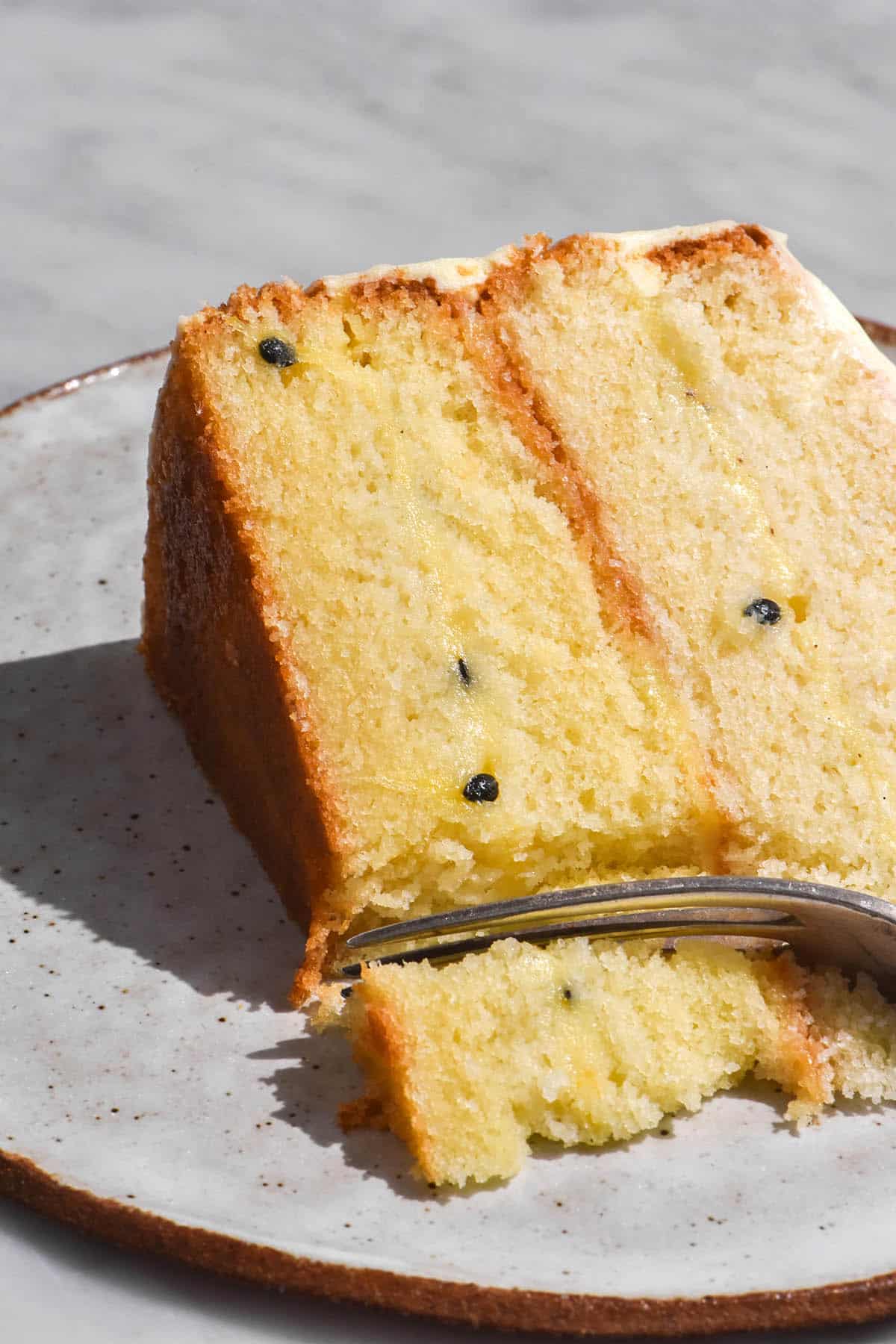 A close up image of a slice of gluten free cake with passion fruit curd filling and cream cheese icing. The slice of cake has a fork sticking into the bottom left corner. It sits on a white ceramic plate atop a white marble table.