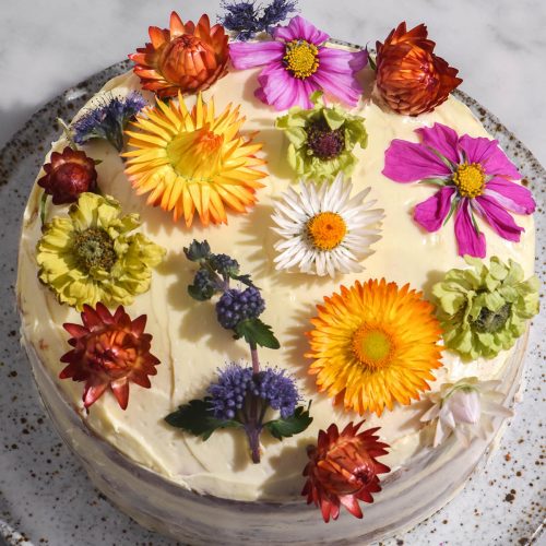 An aerial image of a gluten free cake filled with passion fruit curd and topped with cream cheese icing. The cake is topped with multi coloured flowers and sits on a white speckled ceramic plate atop a white marble table.