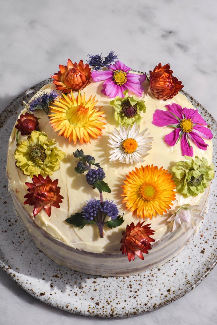 An aerial image of a gluten free cake filled with passion fruit curd and topped with cream cheese icing. The cake is topped with multi coloured flowers and sits on a white speckled ceramic plate atop a white marble table.