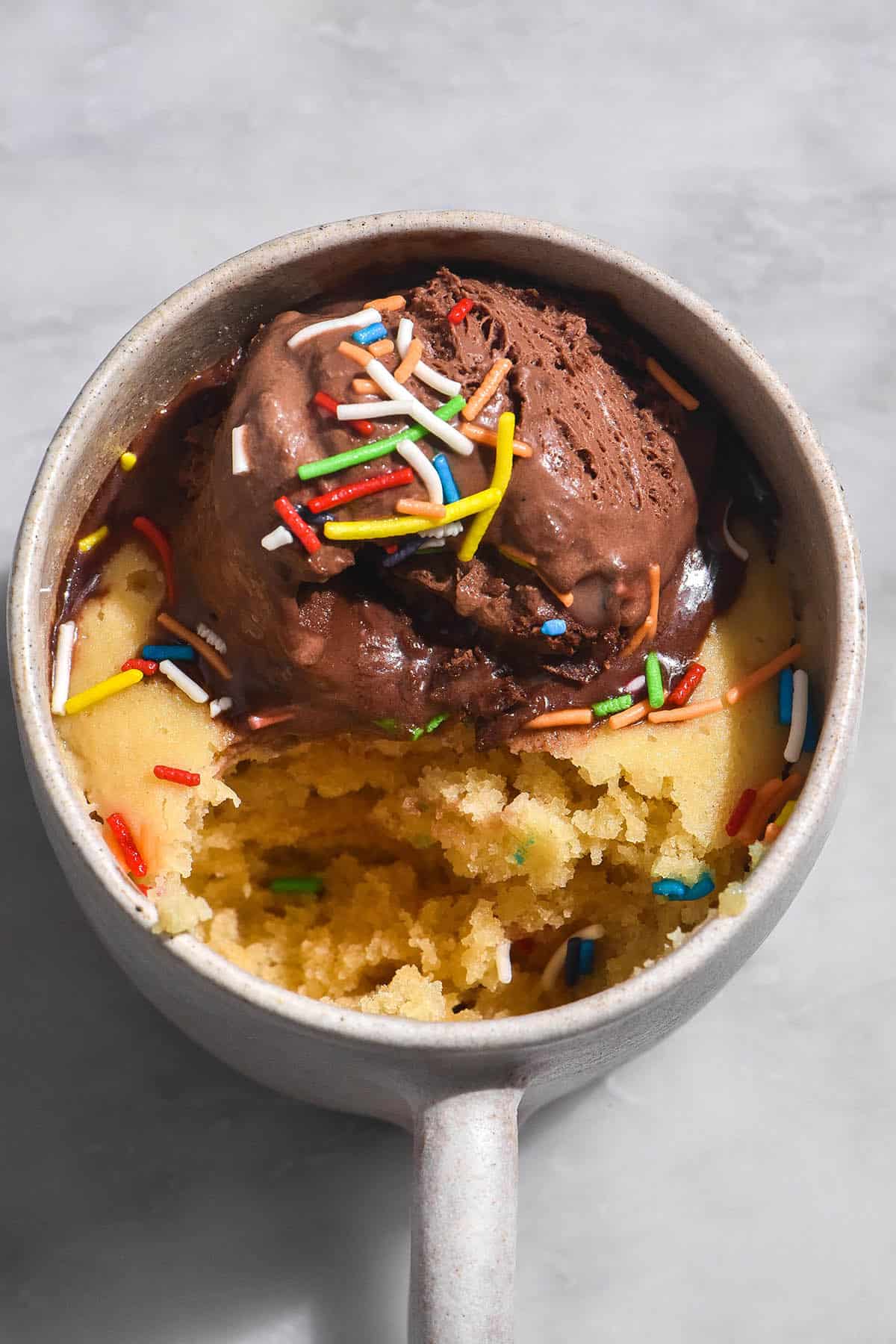An aerial image of a gluten free vanilla mug cake topped with chocolate ice cream and rainbow sprinkles. The mug cake is in a white speckled ceramic mug on a white marble table