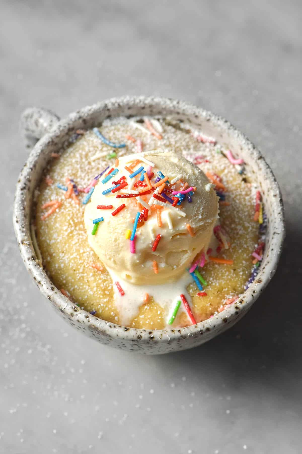 An aerial image of a gluten free vanilla mug cake topped with vanilla ice cream and rainbow sprinkles. The mug cake is in a white speckled ceramic mug on a white marble table