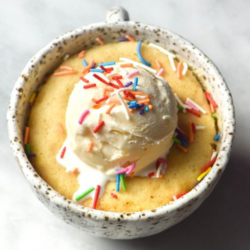 An aerial image of a gluten free vanilla mug cake topped with vanilla ice cream and rainbow sprinkles. The mug cake is in a white speckled ceramic mug on a white marble table