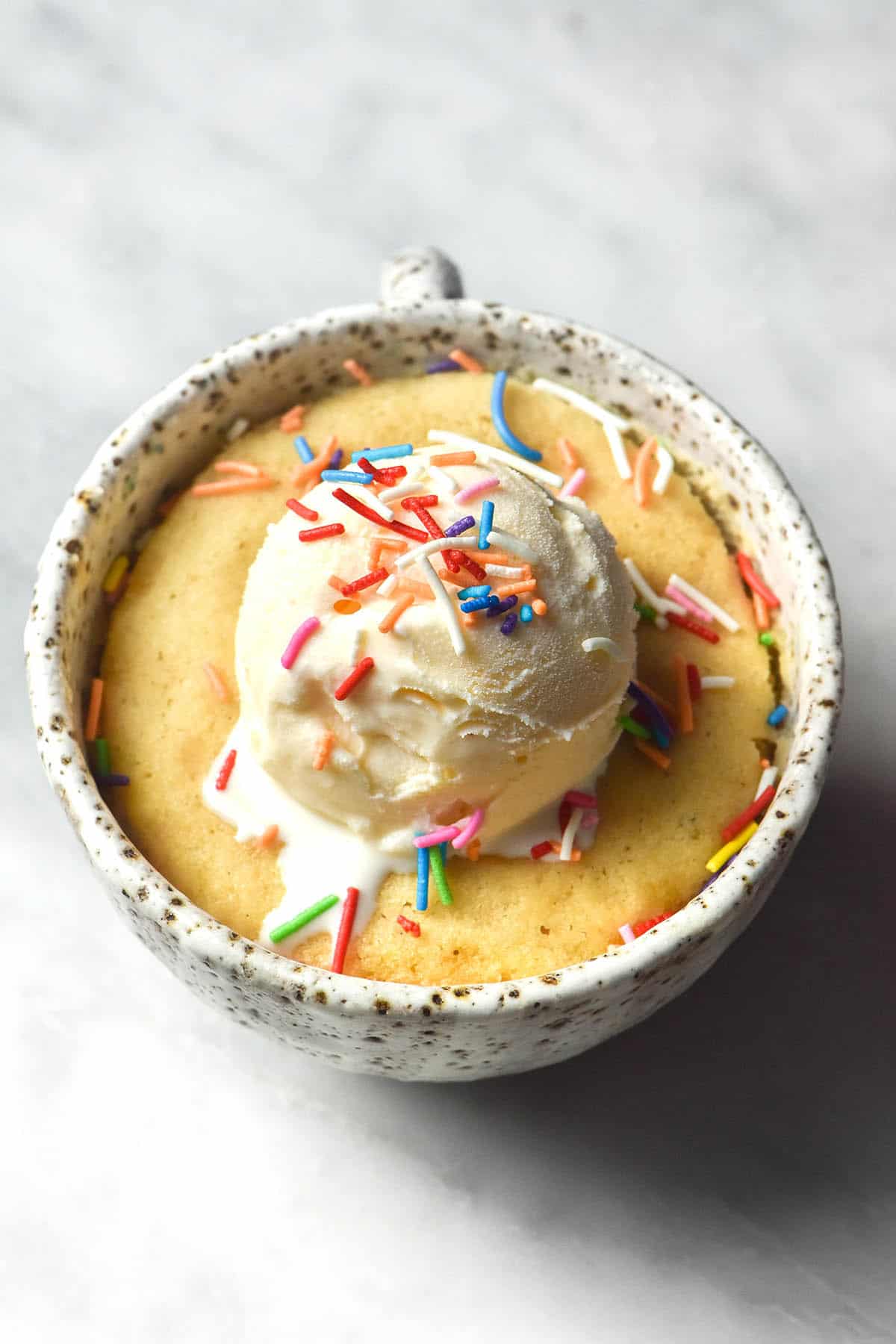 An aerial image of a gluten free vanilla mug cake topped with vanilla ice cream and rainbow sprinkles. The mug cake is in a white speckled ceramic mug on a white marble table