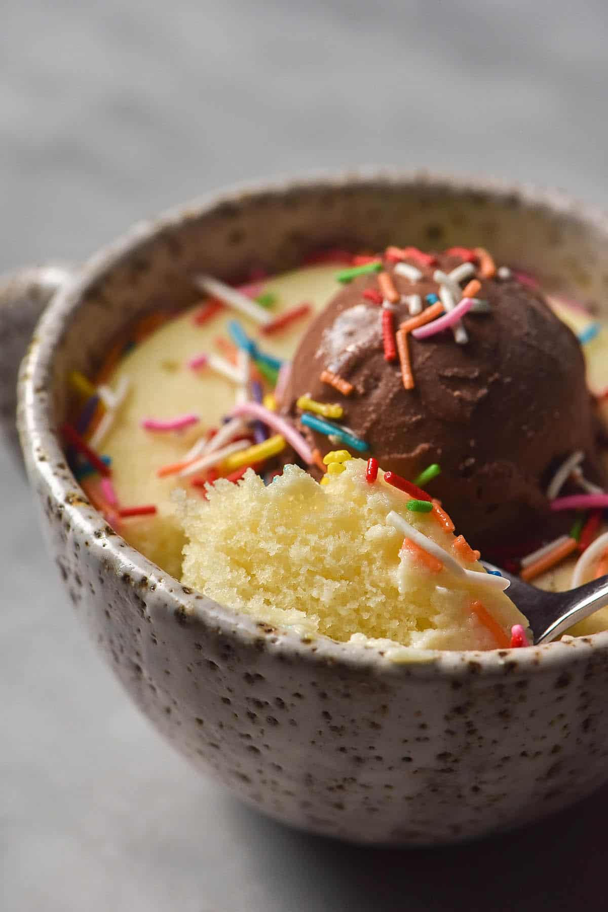 A close up image of the crumb of a gluten free vanilla mug cake. The cake sits in a white speckled ceramic mug on a white marble table. It is topped with rainbow sprinkles and chocolate ice cream. 
