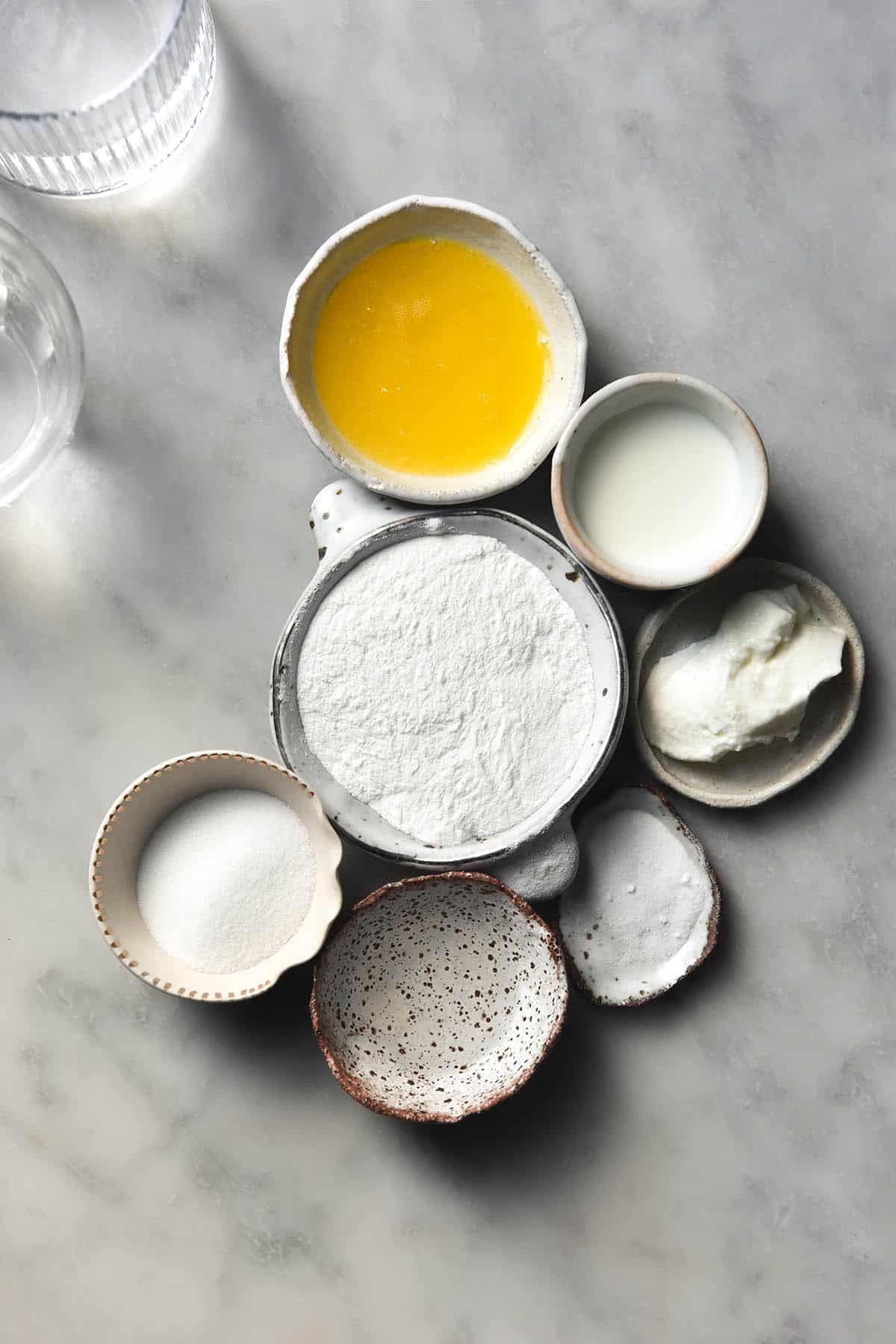 An aerial image of the ingredients used for a gluten free vanilla mug cake. The ingredients are arranged in small white bowls on a white marble table. 