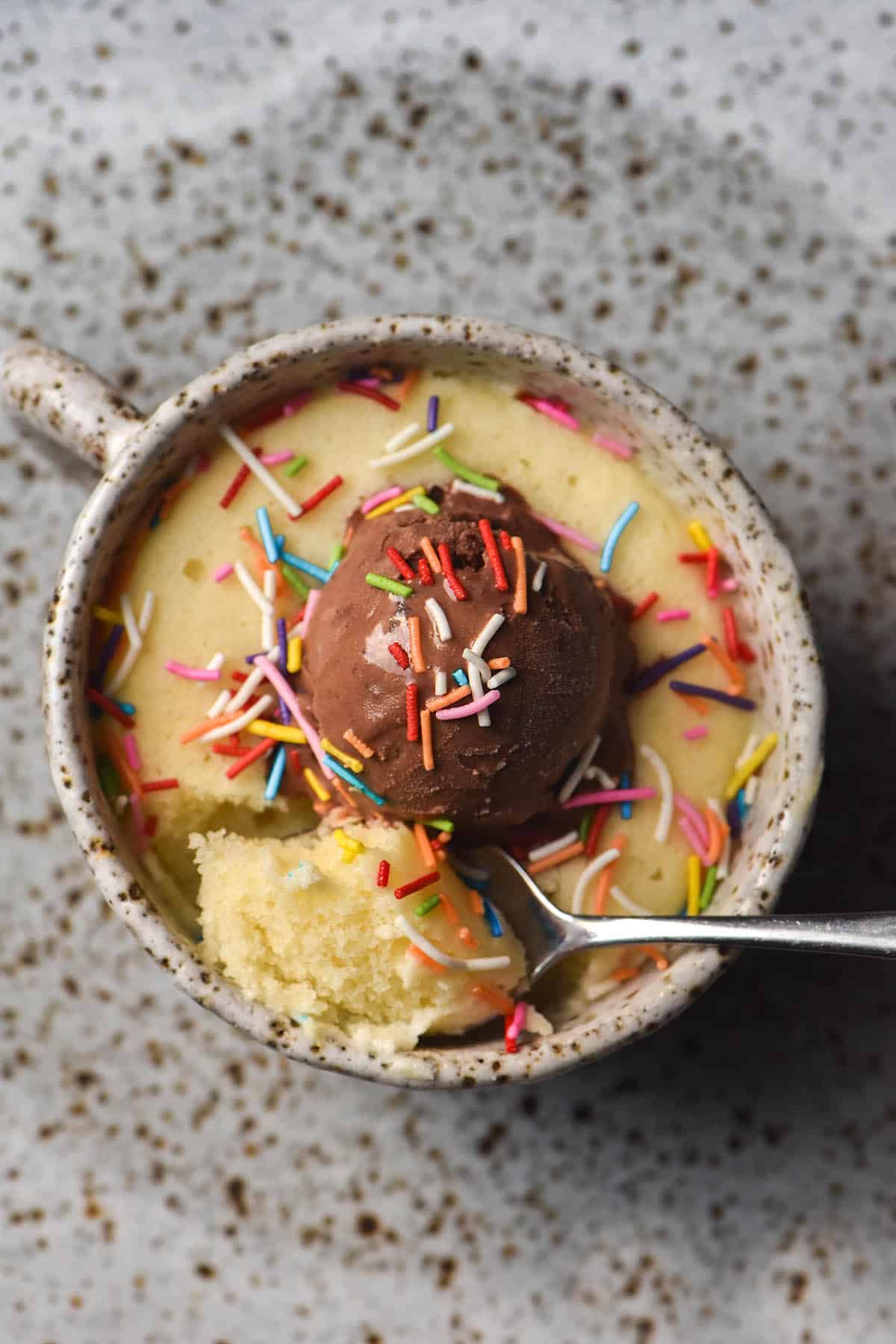 An aerial view of a gluten free mug cake in a white speckled ceramic mug on a white speckled ceramic plate. The mug cake is topped with chocolate ice cream and rainbow sprinkles, and a spoon extends out of the bottom left hand corner. 
