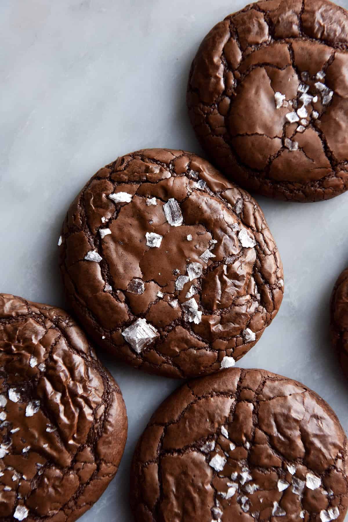 An aerial close up image of gluten free brownie cookies topped with sea salt flakes on a white marble table.