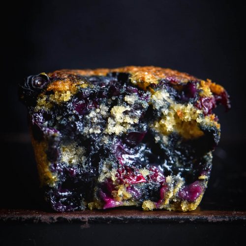 A side on moody macro image of a gluten free vegan blueberry muffin against a black backdrop. The muffin has been torn in half, revealing the blueberry laden muffin crumb inside