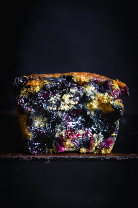 A side on moody macro image of a gluten free vegan blueberry muffin against a black backdrop. The muffin has been torn in half, revealing the blueberry laden muffin crumb inside