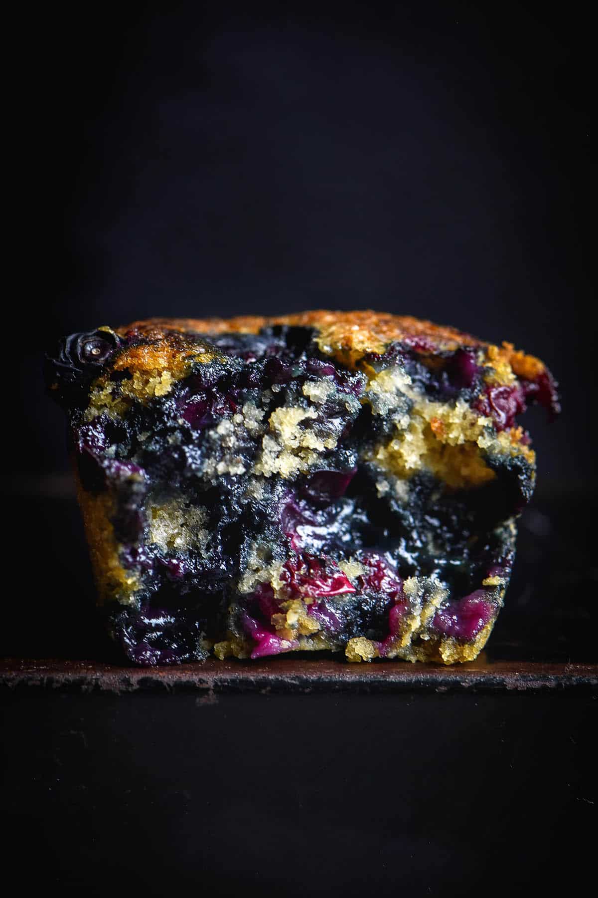 A moody macro image of a gluten free vegan blueberry muffin on a black rustic table against a black background. The muffin has been torn in half, revealing the deeply blueberry coloured crumb inside. 