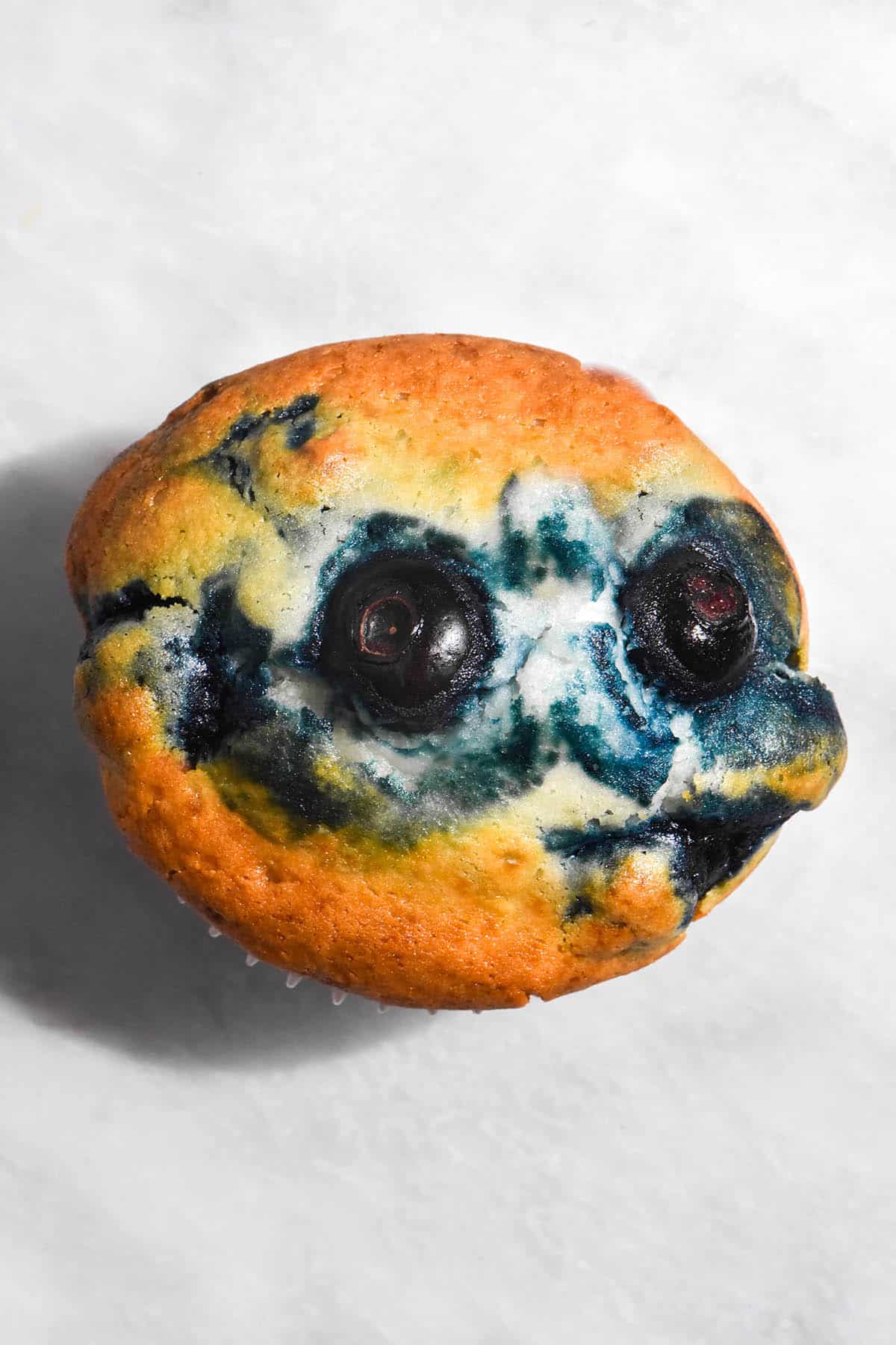 An aerial image of a single serve gluten free vegan blueberry muffin atop a white marble table. The muffin is golden brown and has swirls of blueberry on top.