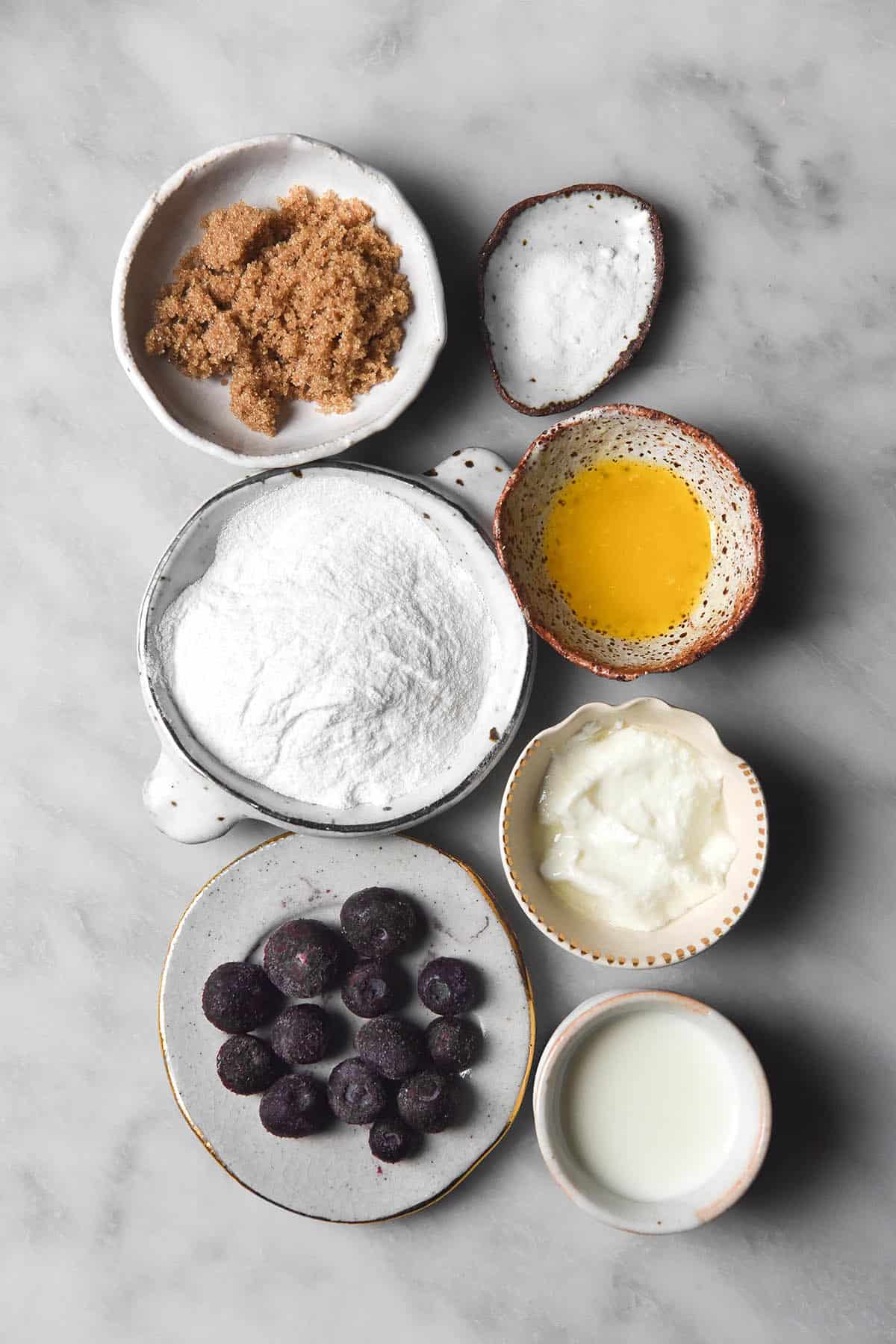 An aerial image of the ingredients needed to make a gluten free vegan blueberry muffin arranged in small white ceramic bowls on a white marble table. 