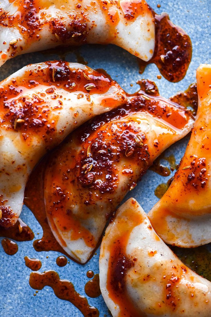 An aerial close up image of gluten free vegan dumplings on a bright blue ceramic plate. The dumplings are smothered in a bright red chilli oil.