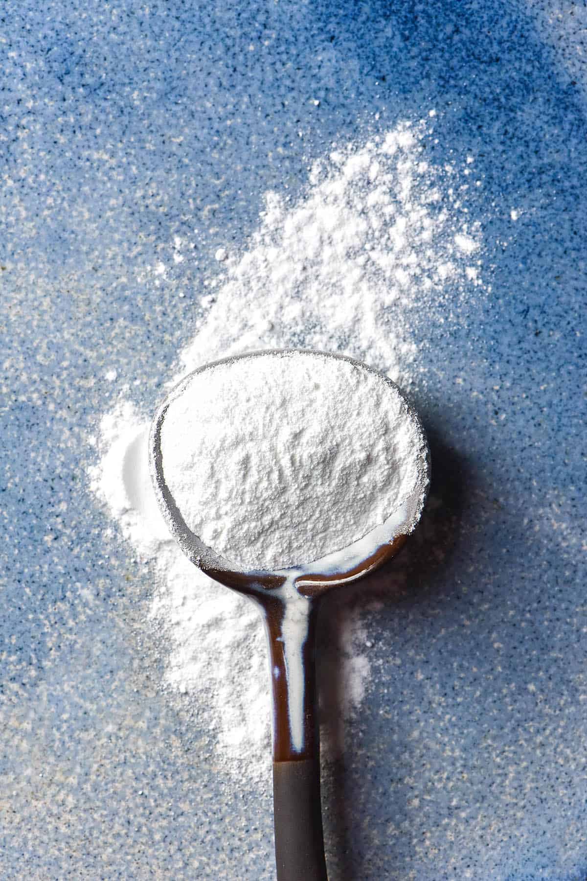 An aerial image of a ceramic spoon filled with gluten free baking powder on a sky blue mottled ceramic plate