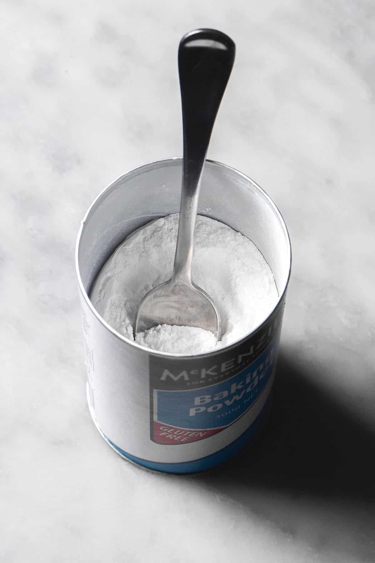 An aerial image of a teaspoon sticking out of an open tub of gluten free baking powder on a white marble table. 