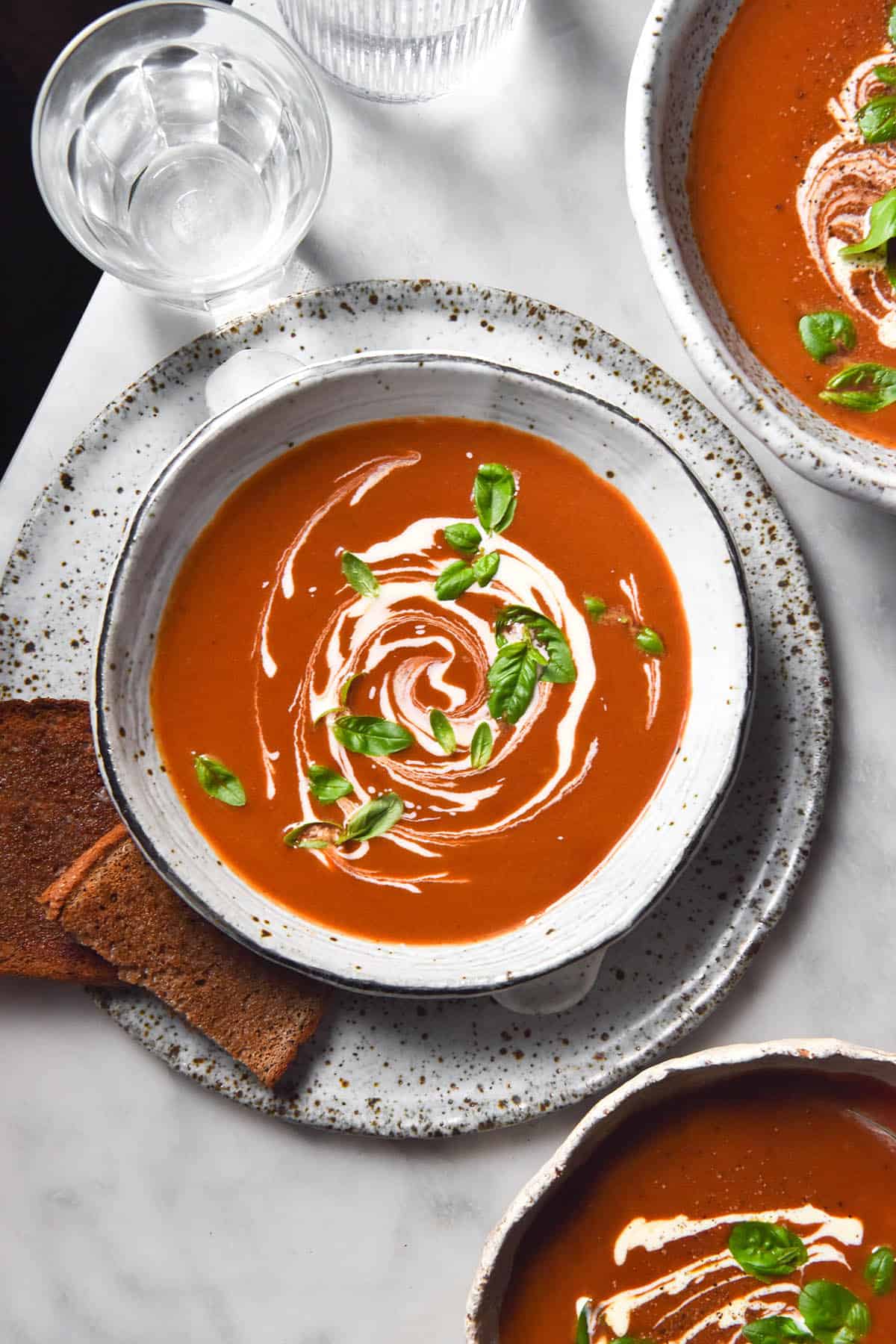 An aerial image of three white ceramic bowls filled with low FODMAP tomato soup on a white marble table. The soup is topped with a swirl of cream, fresh basil leaves and freshly cracked pepper. The central bowl sits on a white speckled ceramic plate with toast sitting to the left of the image. 