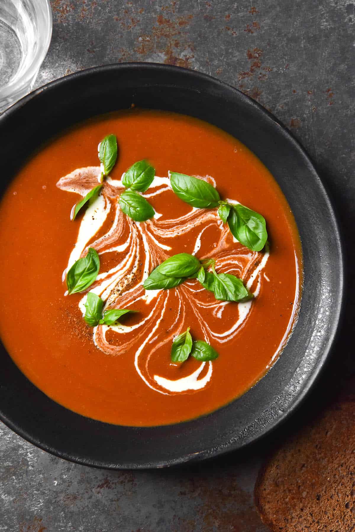 A moody image of a dark ceramic bowl filled with low FODMAP tomato soup. The soup is topped with swirls of cream, fresh basil leaves and freshly cracked pepper. 