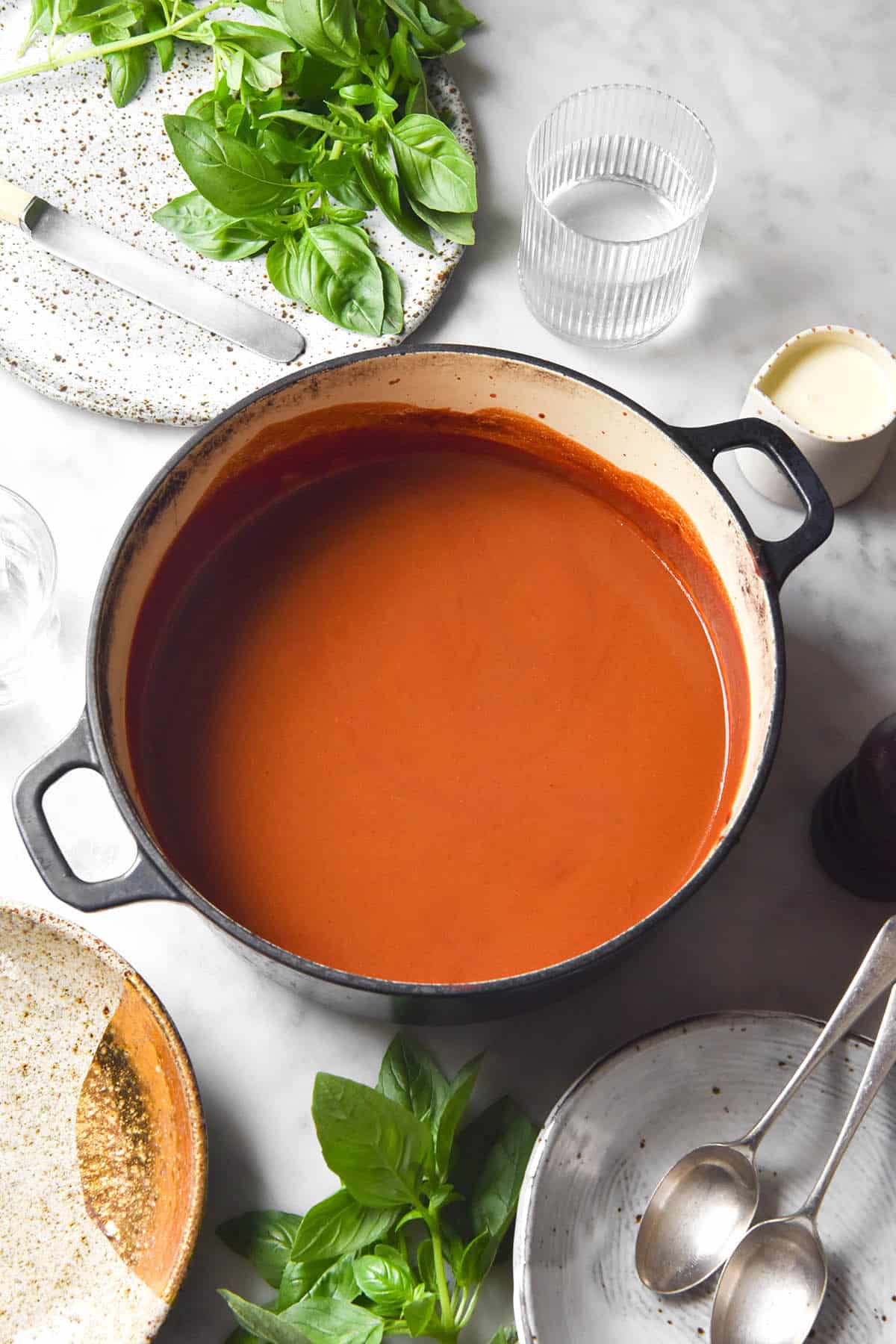 An side on image of a Dutch oven filled with low FODMAP tomato soup. The pot sits atop a white marble table and is surrounded by a white speckled ceramic plate, glasses of water, cream and fresh basil leaves. 