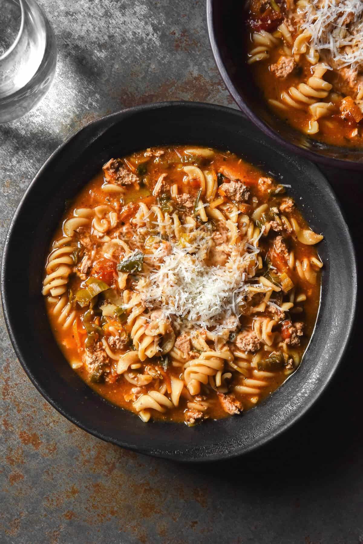 An aerial image of a dark ceramic bowl filled with low FODMAP vegetarian lasagne soup topped with freshly grated parmesan. The bowl sits on a dark metallic backdrop and a second bowl sits in the top right hand corner. 