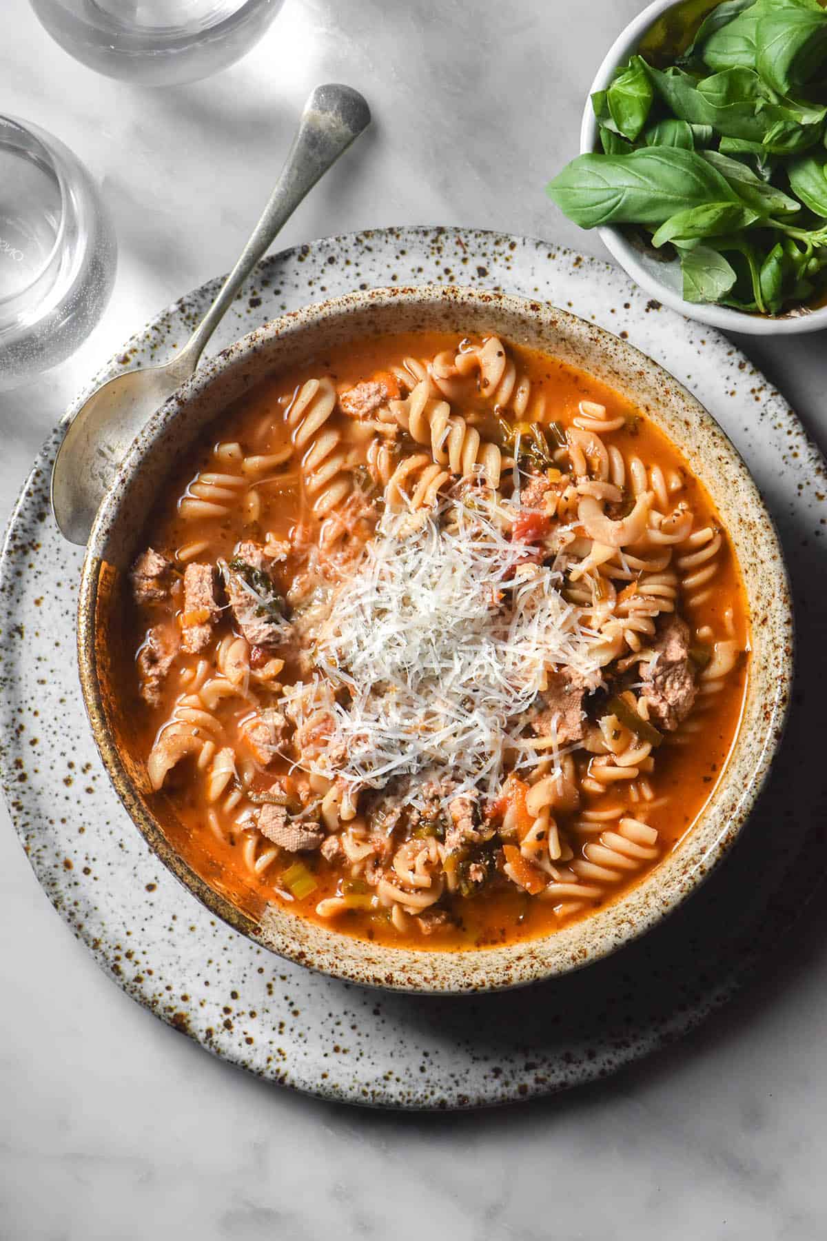 An aerial image of a beige speckled bowl filled with low FODMAP vegetarian lasagne soup atop a white speckled ceramic plate on a white marble table. A small glass of water sits to the left hand side and a bowl of basil sits to the right hand side. 