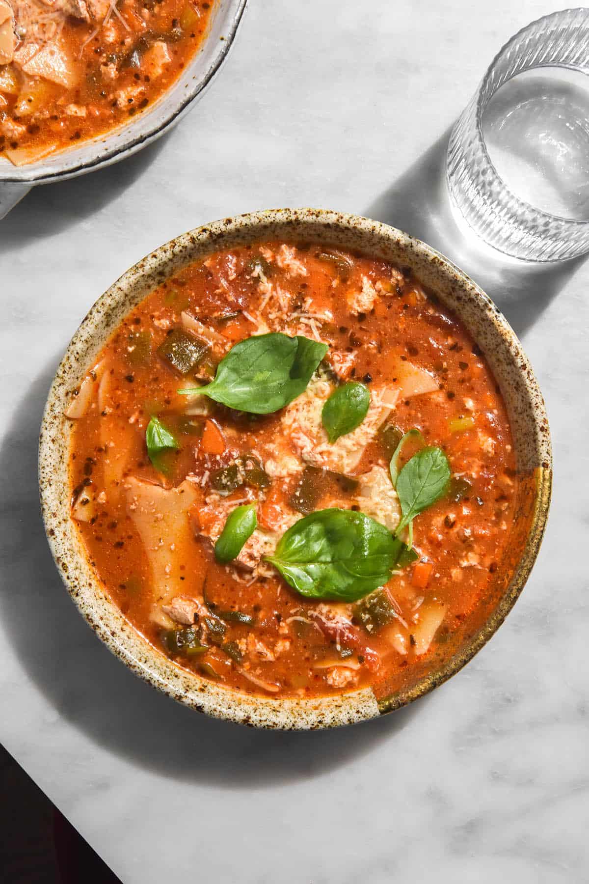 An aerial image of a bowl of low FODMAP vegetarian lasagne soup on a white marble table. The soup is a deep red colour and topped with freshly grated parmesan and basil leaves. 