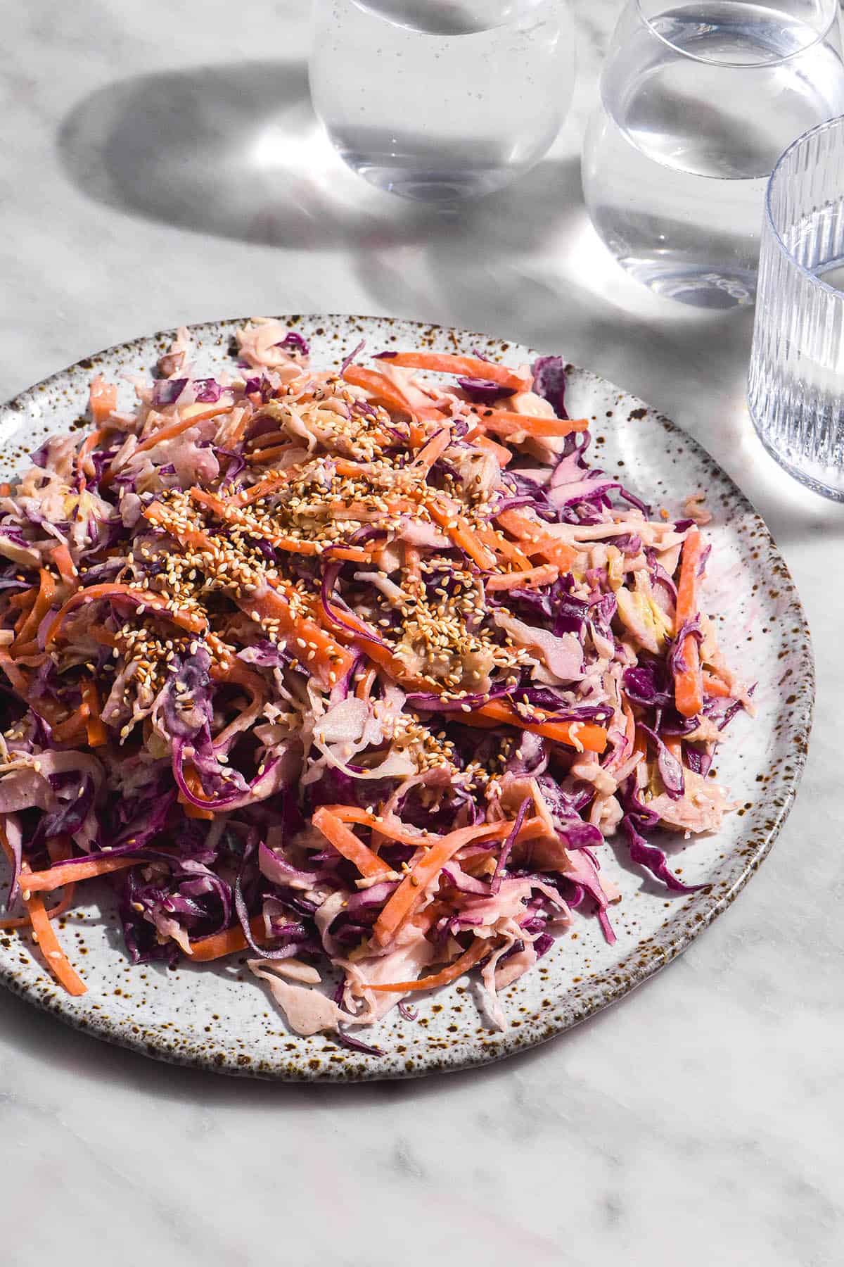 A side on, brightly lit image of a vibrant tahini coleslaw on a white speckled ceramic plate atop a white marble table. 