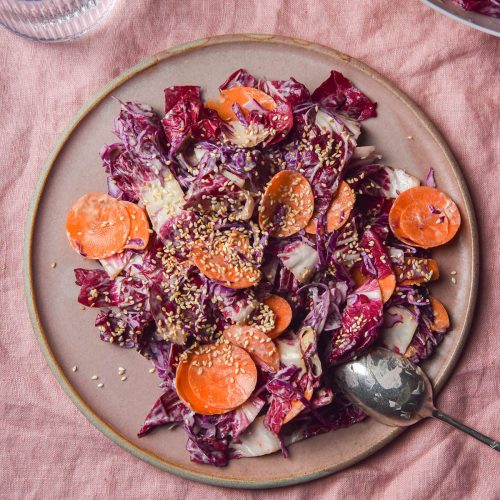 An aerial image of a pale pink ceramic plate topped with tahini coleslaw. The plate sits atop a pale pink linen tablecloth.