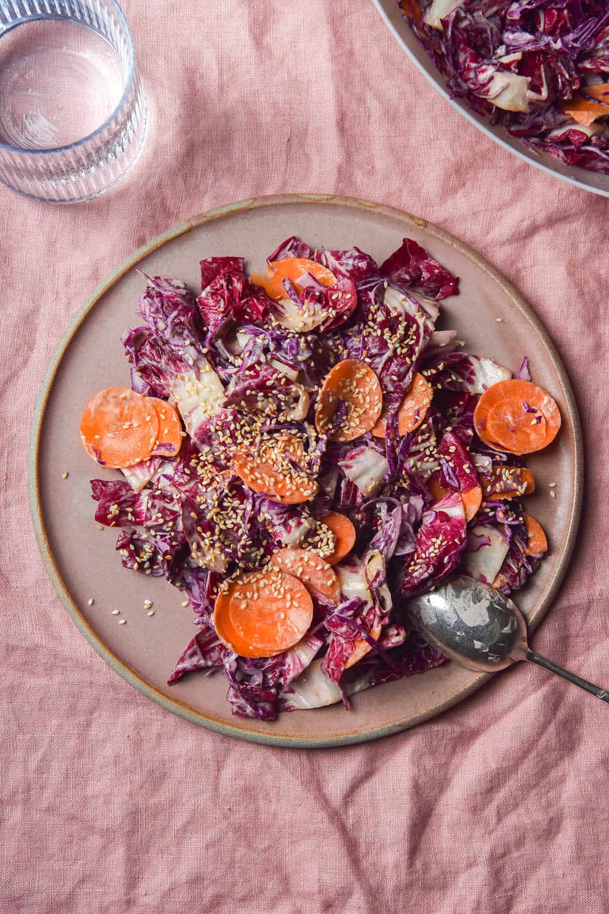 An aerial image of a pale pink ceramic plate topped with tahini coleslaw. The plate sits atop a pale pink linen tablecloth.