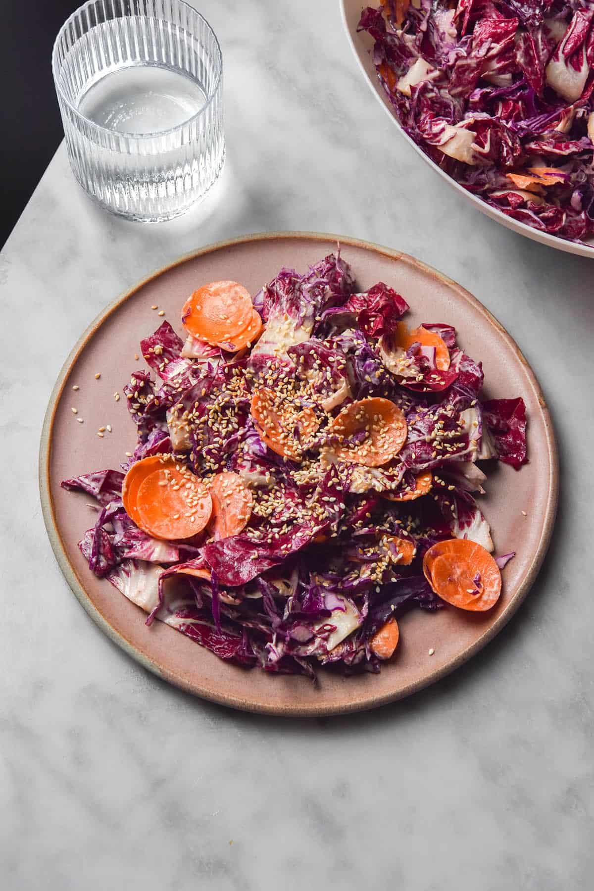 An aerial image of a pale pink ceramic plate topped with tahini coleslaw on a white marble table. A bowl of coleslaw sits in the top right corner while a glass of water sits in the top left corner. 