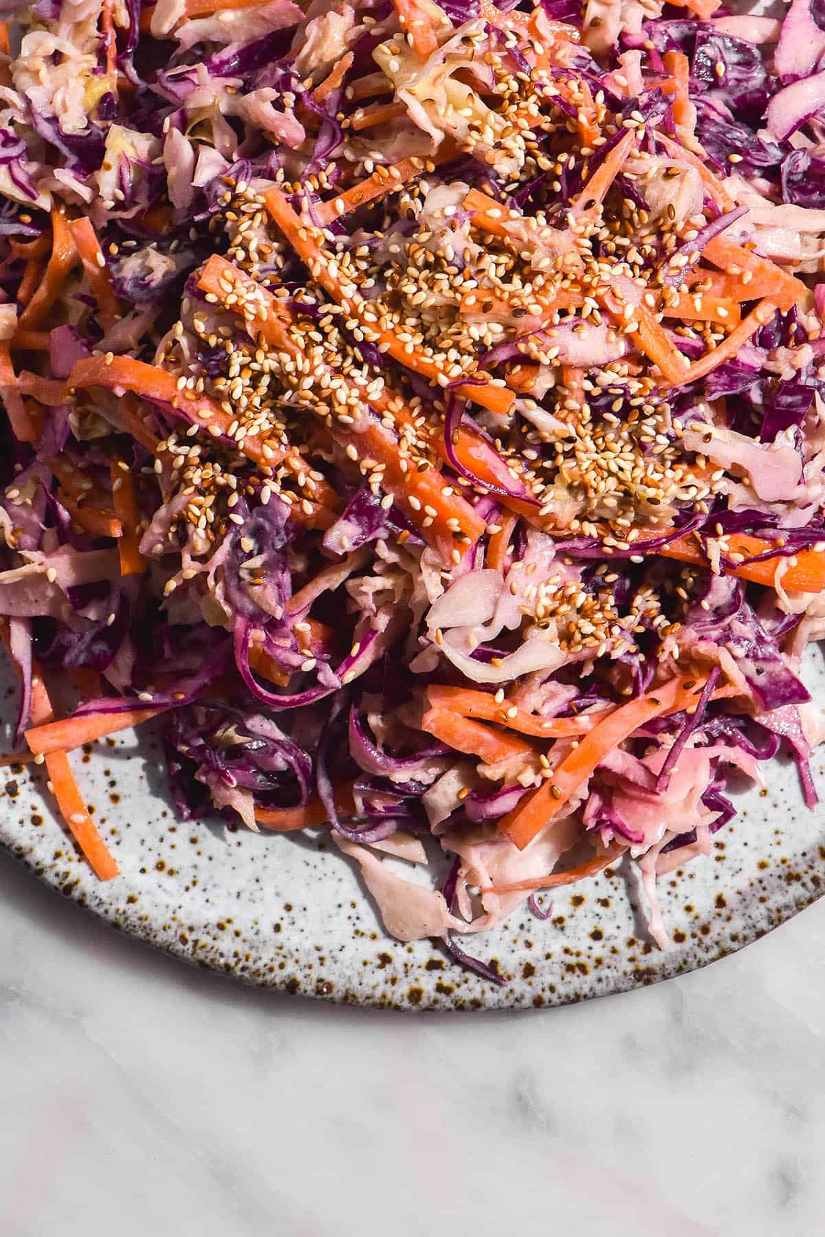 A brightly lit aerial image of a vibrant tahini coleslaw on a white speckled ceramic plate topped with lots of toasted sesame seeds. 