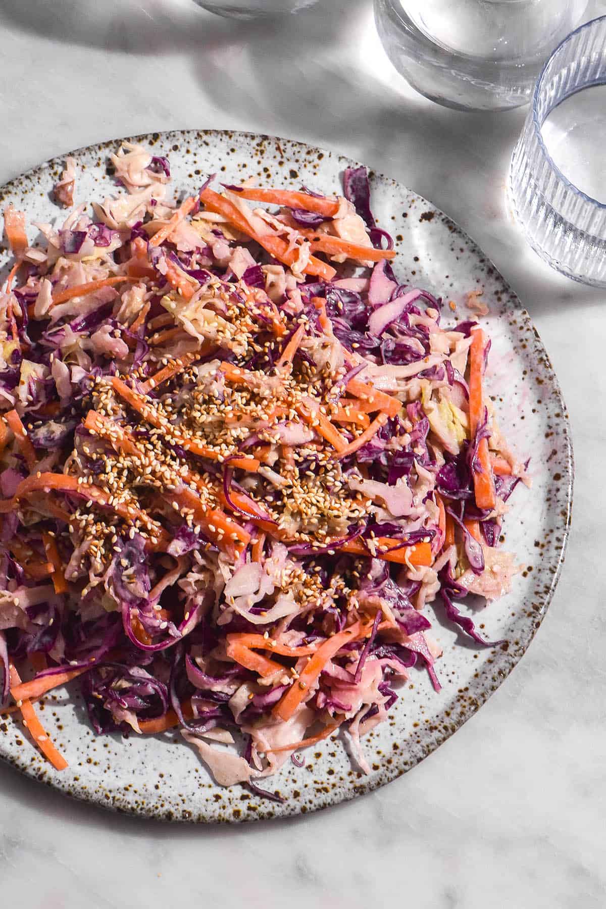 A brightly lit aerial image of a tahini slaw on a white ceramic plate atop a white marble table. The slaw is vibrantly coloured and topped with lots of toasted sesame seeds. 