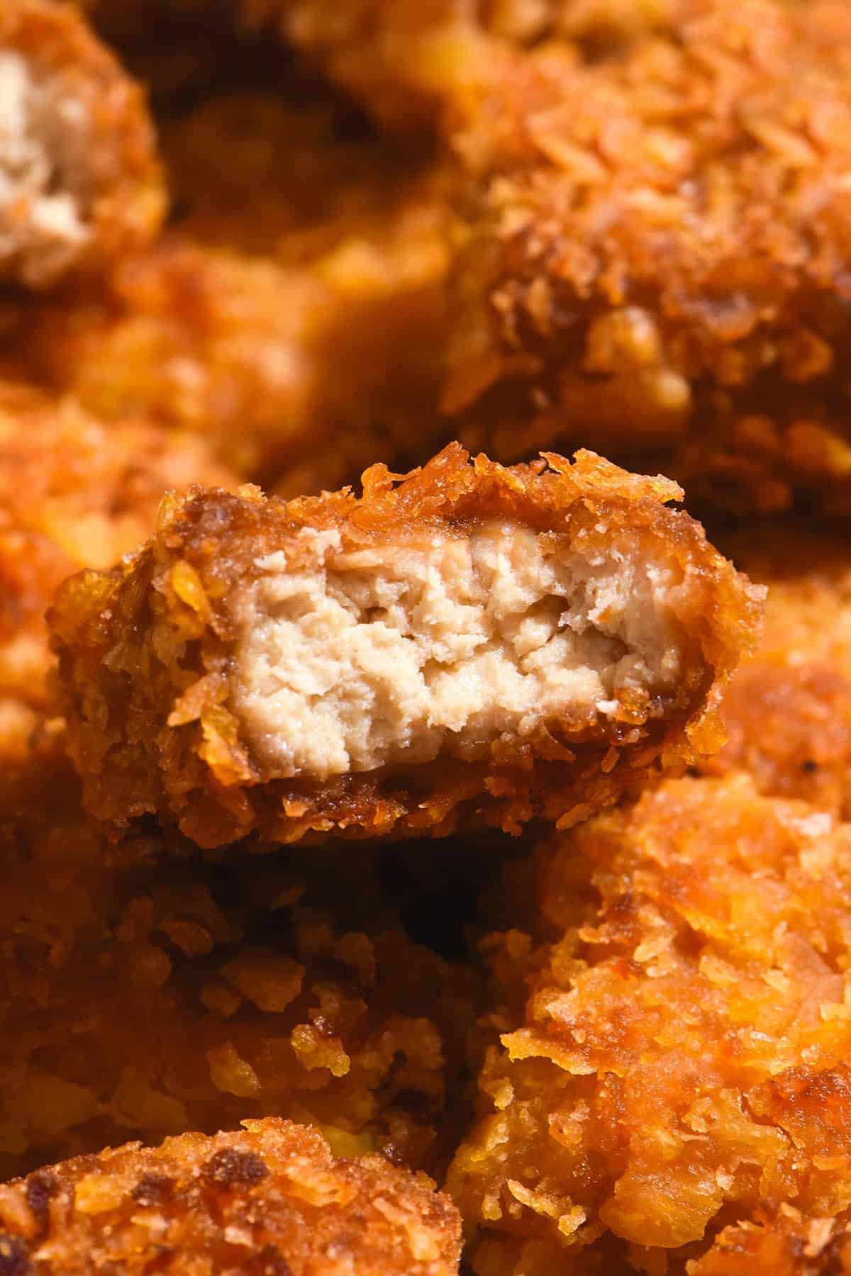 A macro close up image of gluten free tofu nuggets. The central nugget has been bitten into, revealing the juicy 'meat' centre and golden brown crumb