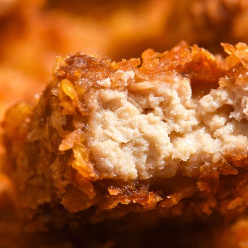 A macro close up image of gluten free tofu nuggets. The central nugget has been bitten into, revealing the juicy 'meat' centre and golden brown crumb