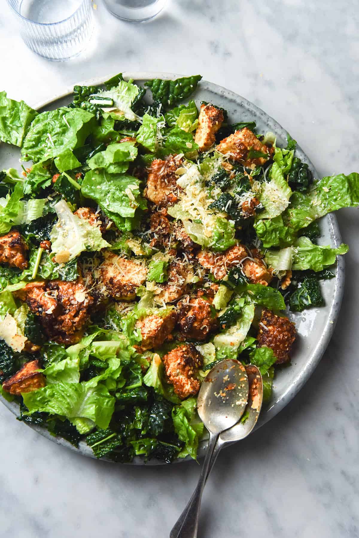 An aerial image of a vegetarian Caesar salad with tofu nuggets on a large white ceramic serving platter atop a white marble table. 