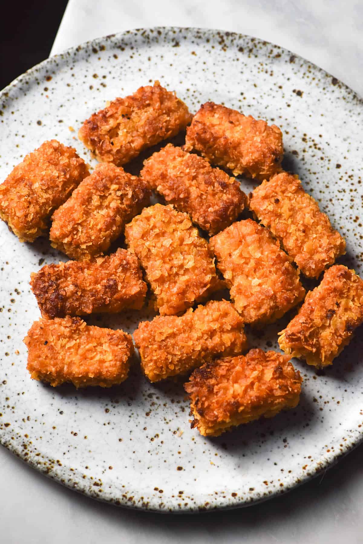 An angled image of gluten free tofu nuggets on a white speckled ceramic plate against a white marble table. 