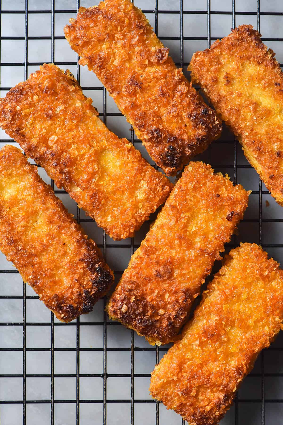 An aerial image of golden brown vegan fish fingers on a wire cooling rack atop a white marble table. 
