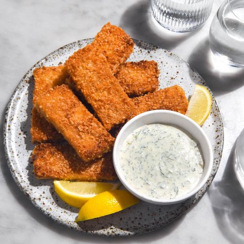 A brightly lit aerial image of low FODMAP vegan tartar sauce in a white bowl surrounded by vegan fish fingers on a white speckled ceramic plate.