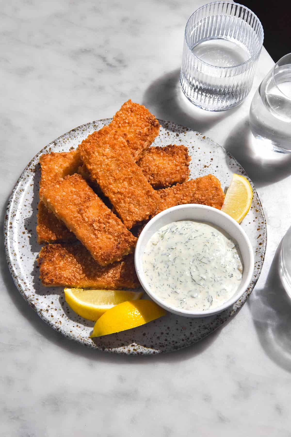 A brightly lit aerial image of low FODMAP vegan tartar sauce in a white bowl surrounded by vegan fish fingers on a white speckled ceramic plate. 