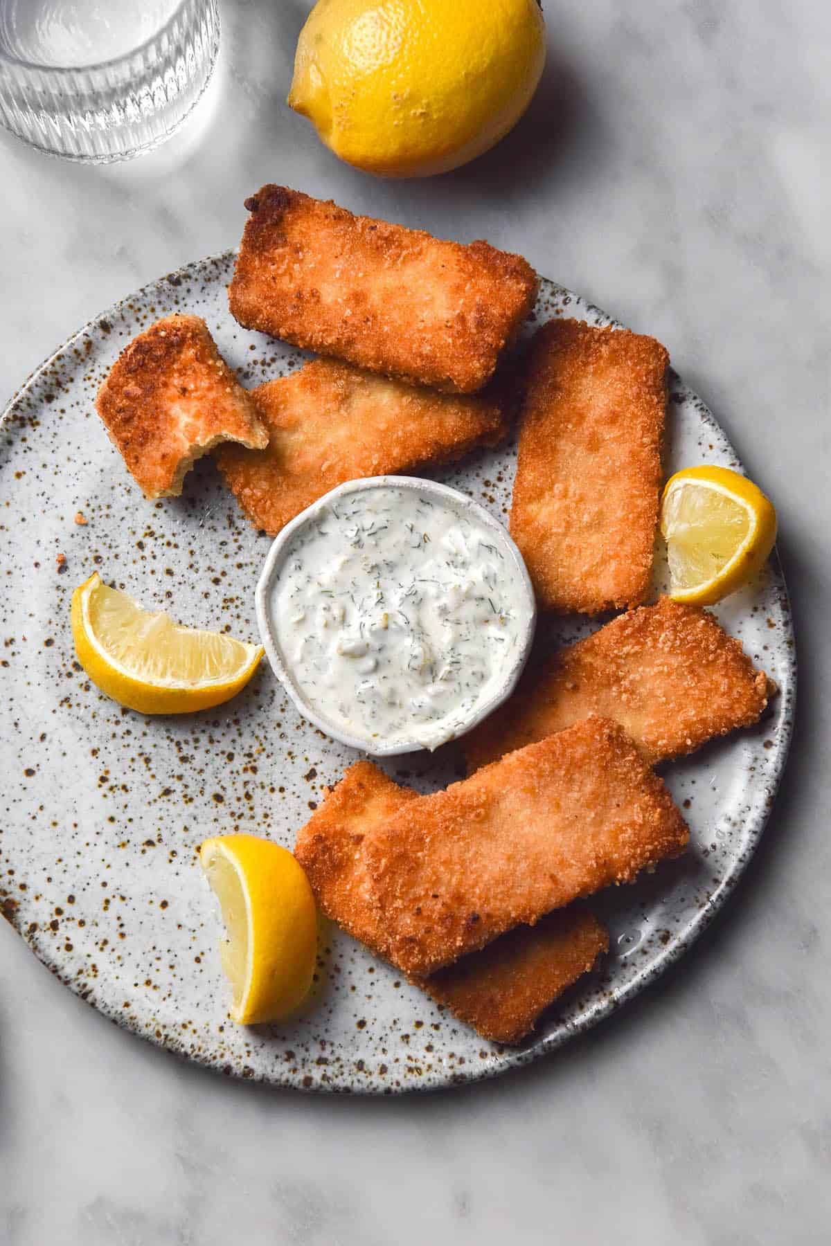 An aerial image of vegan fish fingers casually arranged on a white speckled ceramic plate atop a white marble table. A white bowl of vegan tartar sauce sits in the middle of the plate and three wedges of lemon are strewn amongst the fish fingers. 