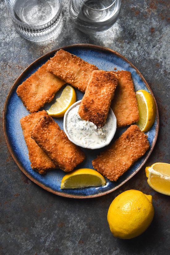 An aerial image of gluten free vegan fish fingers on a bright blue ceramic plate atop a medium blue steel backdrop. A bowl of vegan tartar sauce sits in the middle of the plate and the fish fingers are surrounded by wedges of lemon.