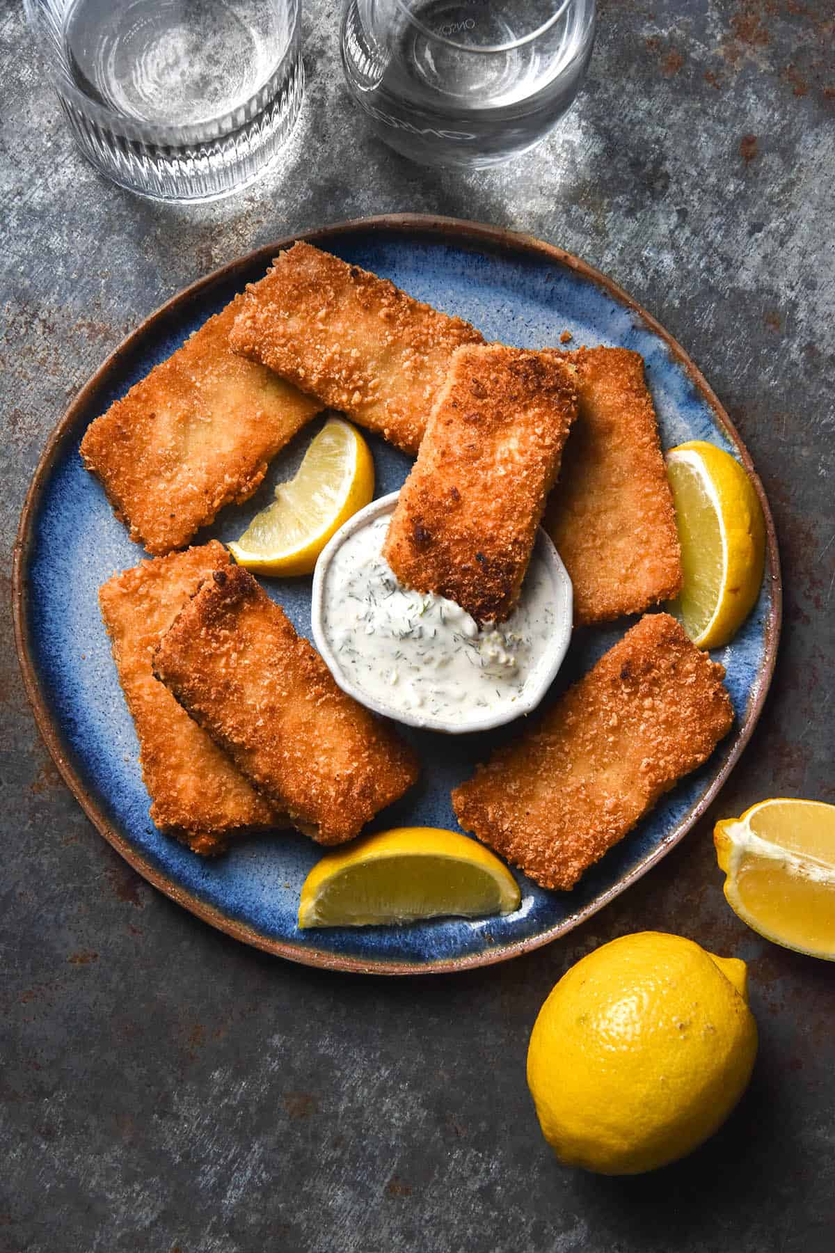 An aerial image of gluten free vegan fish fingers on a bright blue ceramic plate atop a medium blue steel backdrop. A bowl of vegan tartar sauce sits in the middle of the plate and the fish fingers are surrounded by wedges of lemon. 