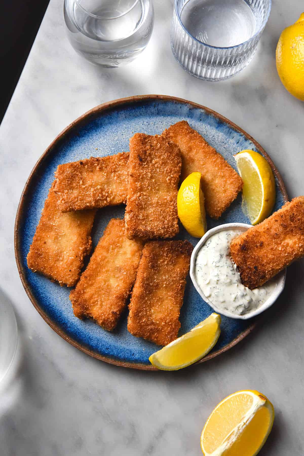 An aerial image of vegan fish fingers casually arranged on a bright blue ceramic plate atop a white marble table. A white bowl of vegan tartar sauce sits in the middle of the plate and three wedges of lemon are strewn amongst the fish fingers. 