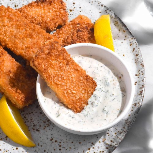 An aerial image of vegan fish fingers casually arranged on a white speckled ceramic plate atop a white marble table. A white bowl of vegan tartar sauce sits in the middle of the plate and three wedges of lemon are strewn amongst the fish fingers.