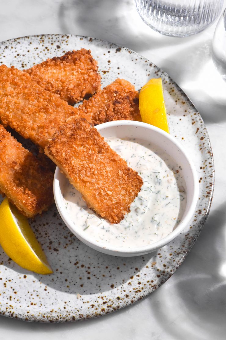 An aerial image of vegan fish fingers casually arranged on a white speckled ceramic plate atop a white marble table. A white bowl of vegan tartar sauce sits in the middle of the plate and three wedges of lemon are strewn amongst the fish fingers.