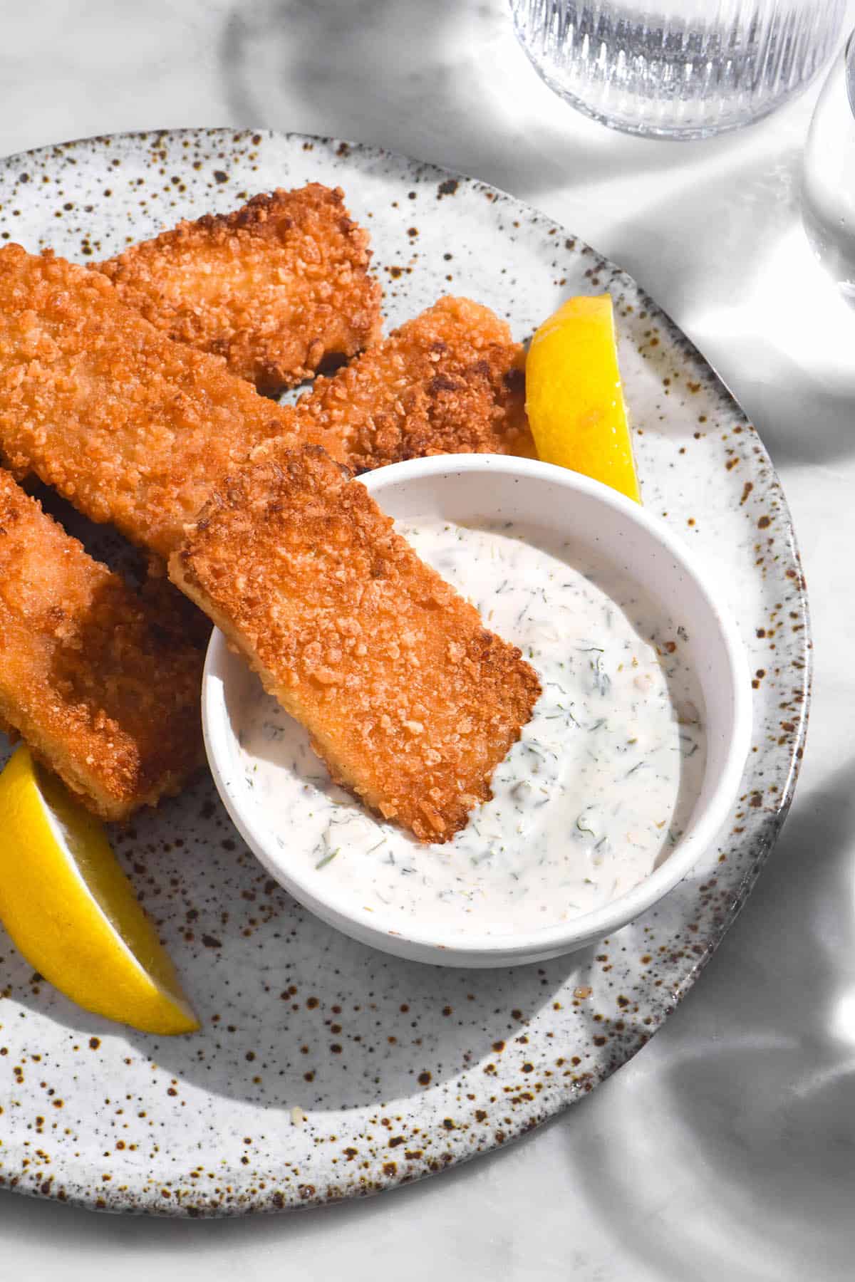 A side on close up image of vegan fish fingers casually arranged on a white speckled ceramic plate atop a white marble table. A white bowl of vegan tartar sauce sits in the middle of the plate and three wedges of lemon are strewn amongst the fish fingers. 