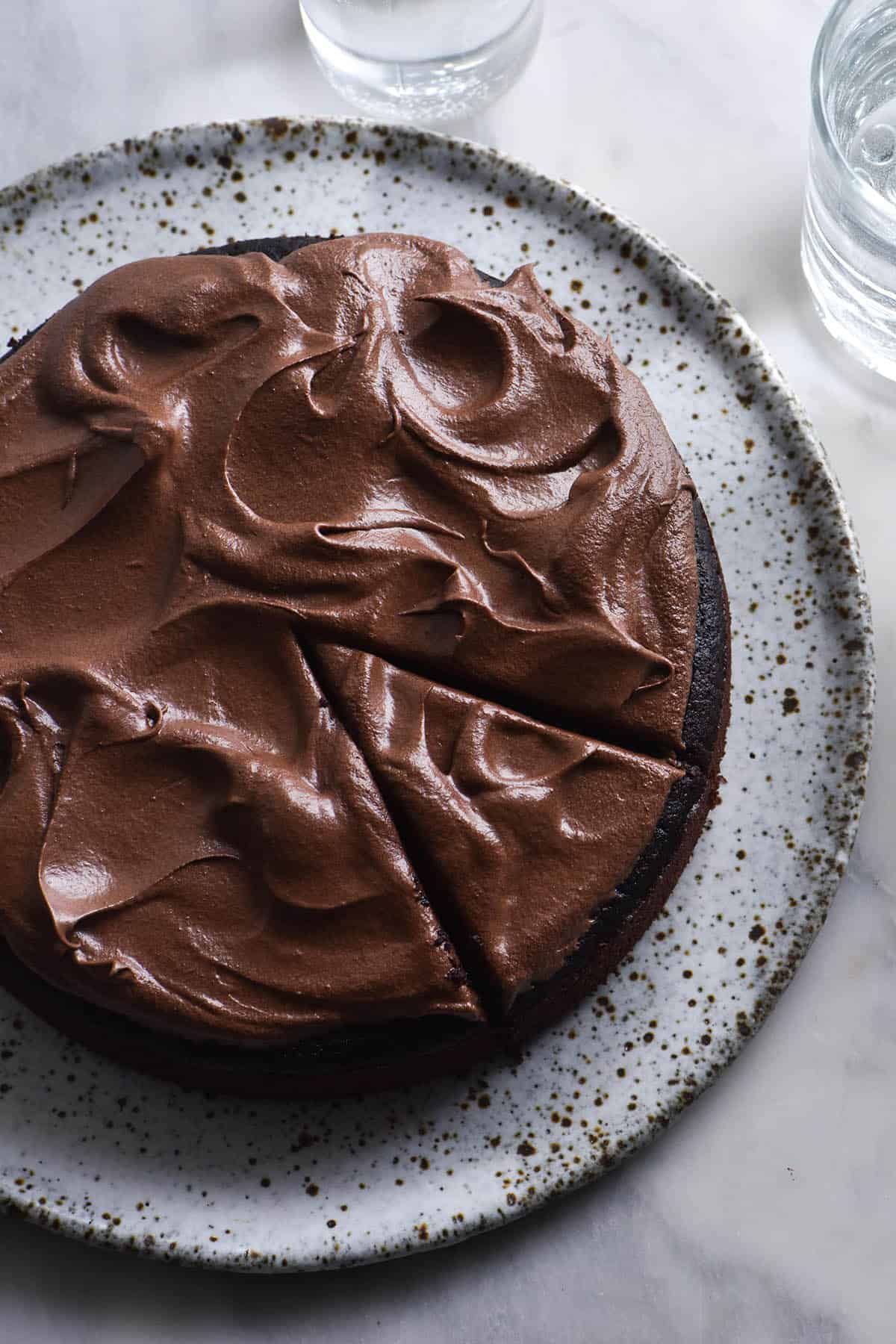 An aerial image of a vegan gluten free chocolate cake on a white speckled ceramic plate atop a white marble table.