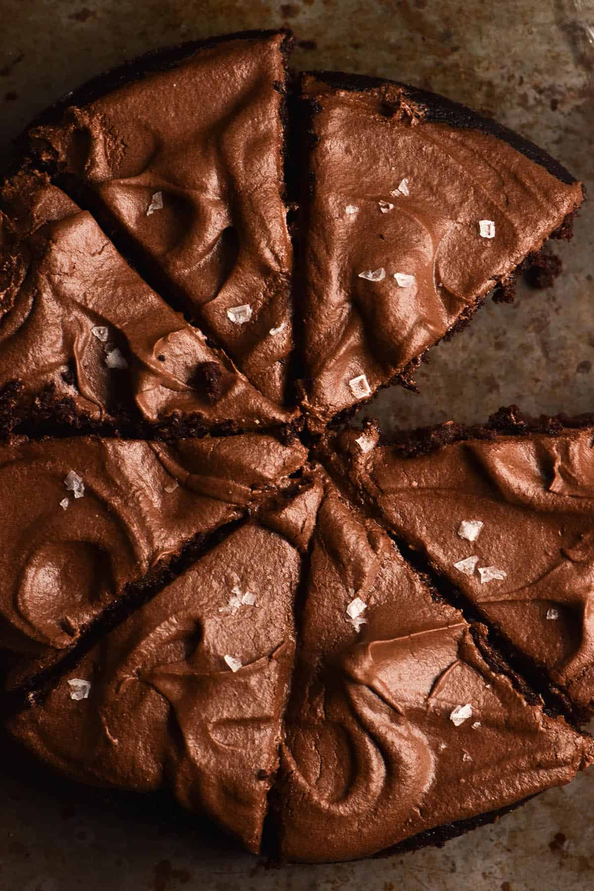 An aerial close up of a vegan gluten free chocolate cake topped with chocolate buttercream and sea salt flakes. The cake has been sliced and one slice has been removed, revealing the dark steel backdrop underneath.