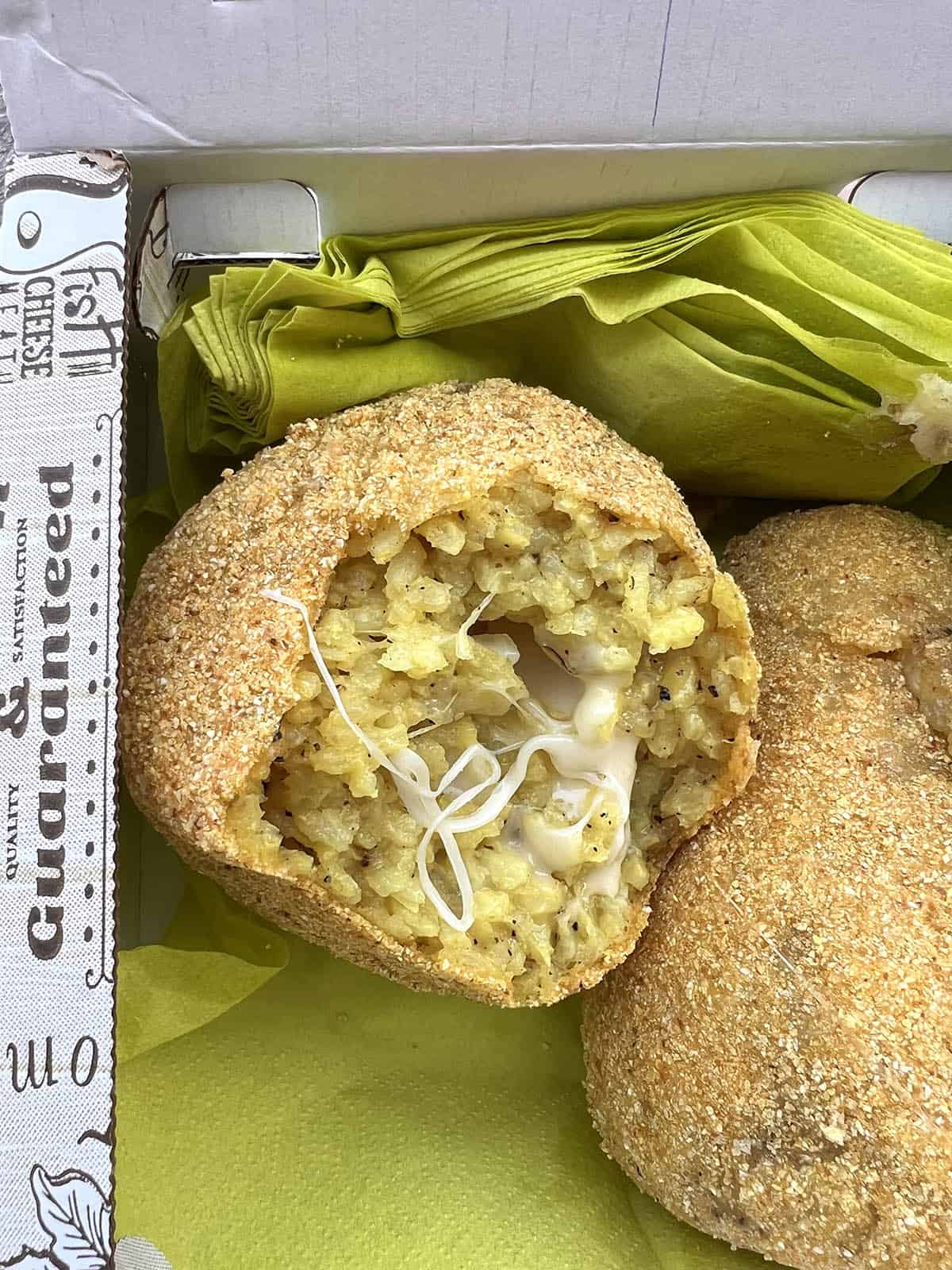 An image of takeaway gluten free arancini in a takeaway box from Arancina in Ortigia. One arancini has been bitten into, revealing the melty cheese inside. 