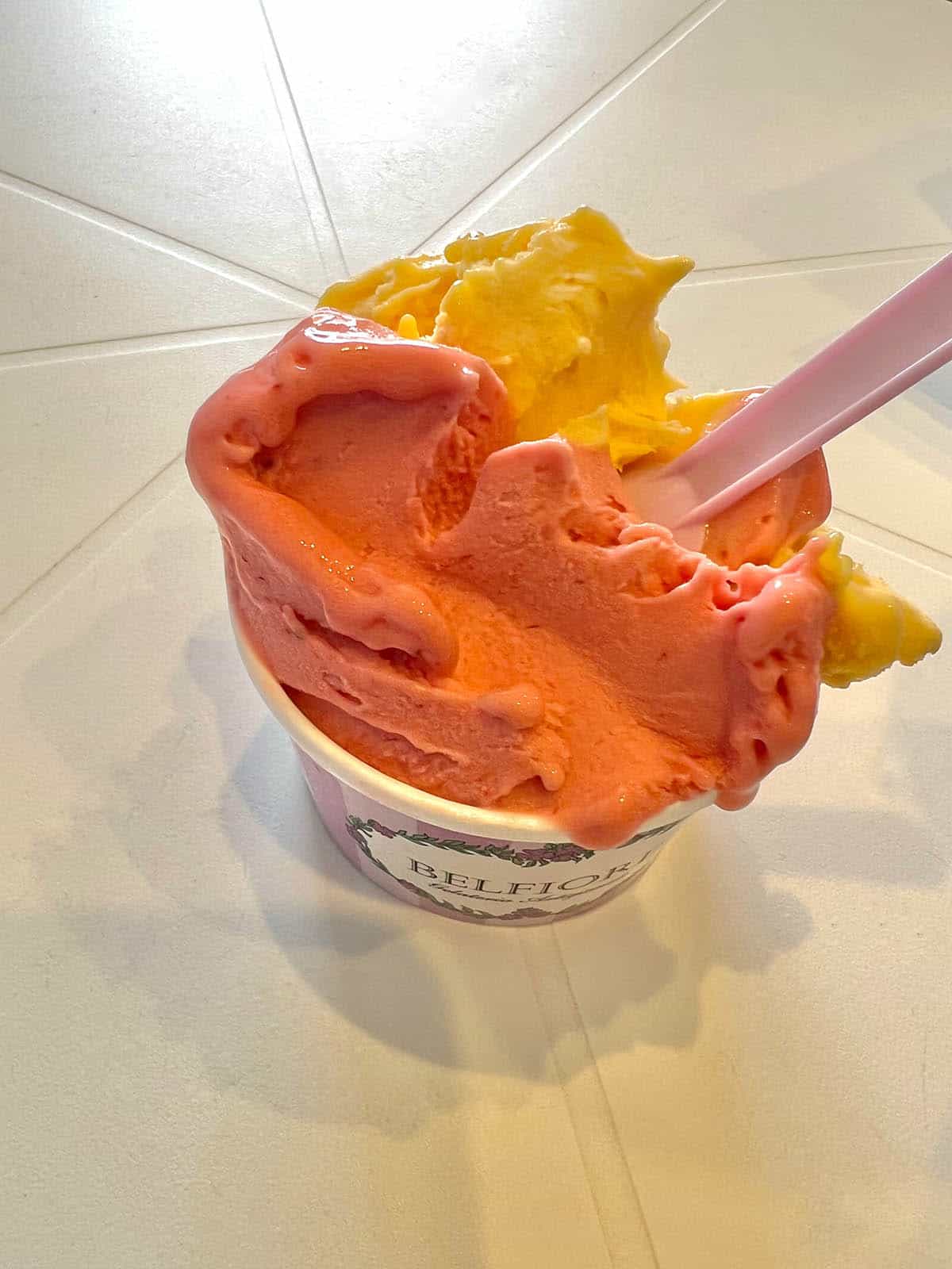 An image of a strawberry and mango cup of gelato on a white plastic table in indoor lighting in Sicily. 